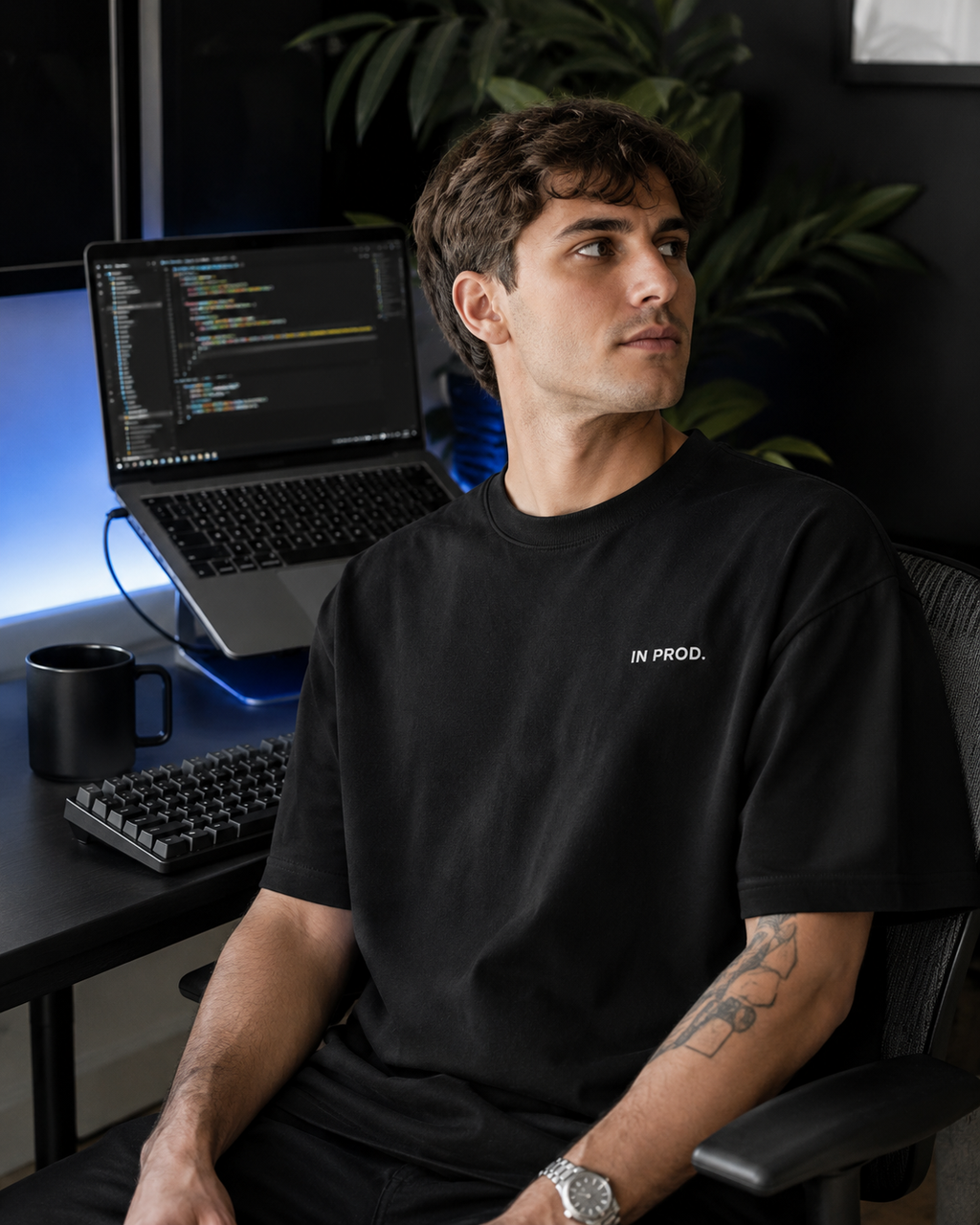 Man sitting at a desk with a laptop and computer setup, wearing a black t-shirt with 'IN PROD.' text.